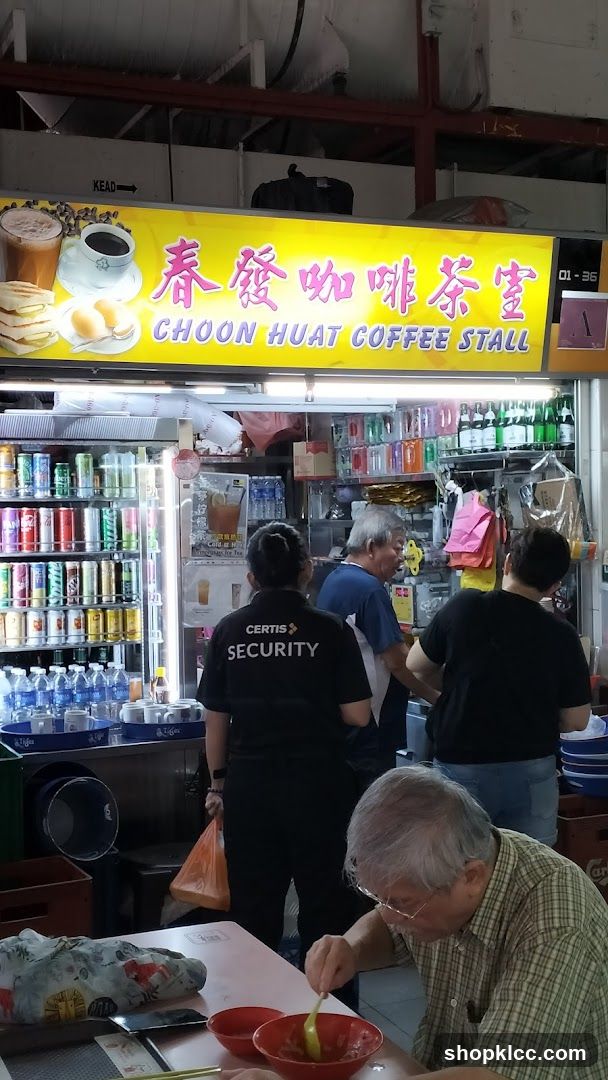 A bright yellow sign with Chinese characters and the words "CHOON HUAT COFFEE STALL" is displayed above a coffee stall. The sign features illustrations of coffee, tea, toast, and boiled eggs. Below the sign, the stall is stocked with various canned drinks and bottled water. Several people are visible, including staff members and customers. One person in the foreground is eating at a table.