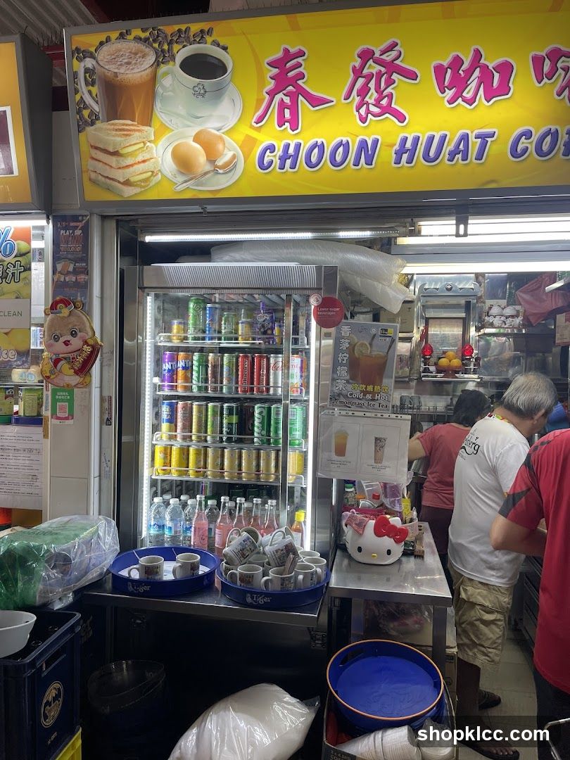 A street-level view of the exterior of Choon Huat Coffee, a food and beverage stall. The shop front features a prominent yellow sign with the business name in Chinese and English, accompanied by illustrations of coffee, toast, and boiled eggs. Below the sign, a glass-fronted refrigerator displays a variety of canned and bottled beverages. In front of the refrigerator, trays of mugs are visible, along with a Hello Kitty-themed dispenser. Two individuals are seen behind the counter, appearing to serve customers.