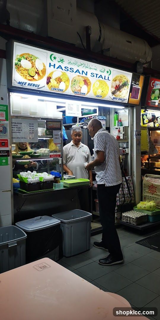 A food stall with a large illuminated sign displaying various dishes like Mee Rebus, Lontong, Mee Siam, Laksa, and Mee Bakso. Two men are interacting at the counter, with one appearing to be a customer and the other an employee. The stall has a self-service and cash-only policy indicated.