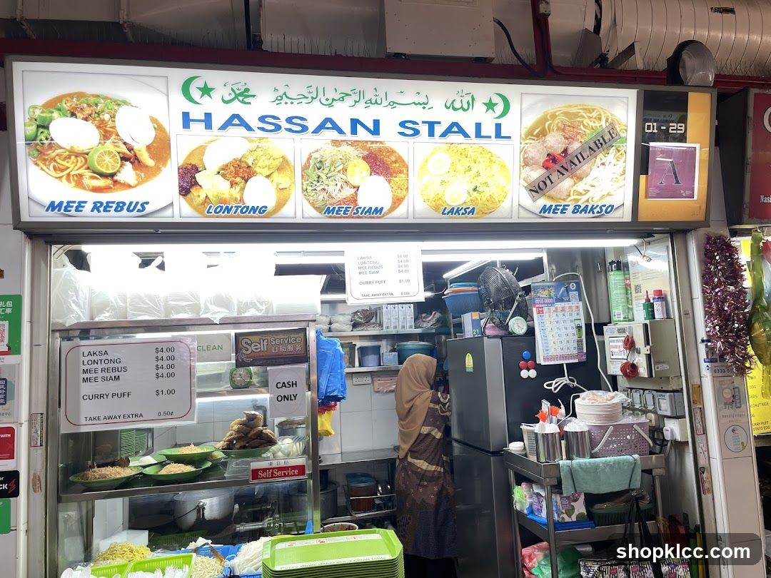 A food stall with a large illuminated sign displaying pictures and names of various dishes. Below the sign is the stall counter with food items displayed and a menu board. A person is visible behind the counter.