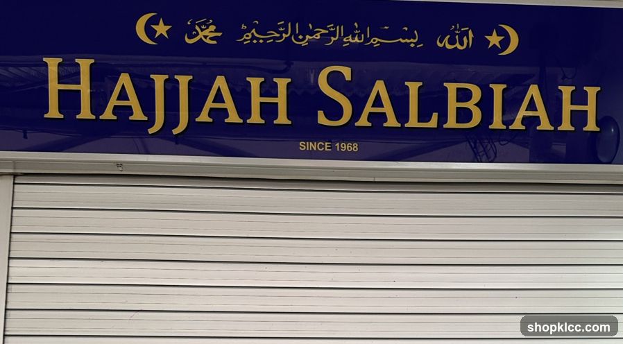 A dark blue sign with gold lettering reads "Hajjah Salbiah" and "SINCE 1968". Above the name, in smaller gold lettering, is Arabic script and a crescent moon with a star. The sign is mounted above a closed, silver, corrugated metal shutter.