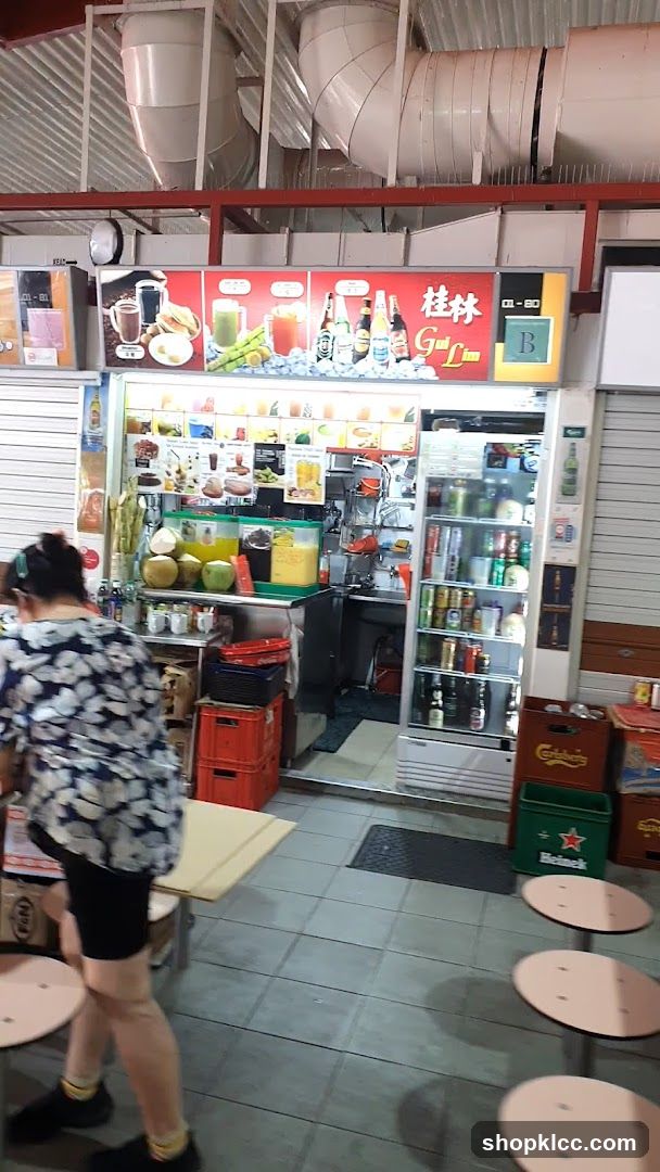 A street-level view of a drink stall with a prominent sign displaying "Gui Lin" in English and Chinese. The stall offers a variety of beverages, with images of drinks and ingredients like sugarcane and coconuts displayed on the menu board. A refrigerator unit is stocked with canned and bottled drinks. The stall appears to be located in an open-air food court or market, with tables and chairs visible in the foreground.
