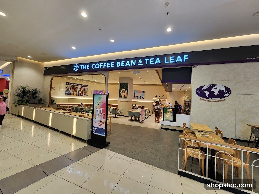 The exterior of a Coffee Bean & Tea Leaf cafe located in a mall. The storefront features the brand's name in large illuminated letters above the entrance. Inside, tables and chairs are visible, along with a counter area. A digital display board with promotional images is placed outside the entrance. A large oval logo with a world map and the text "DESIGN YOUR OWN JOURNEY" is mounted on the wall to the right of the entrance.
