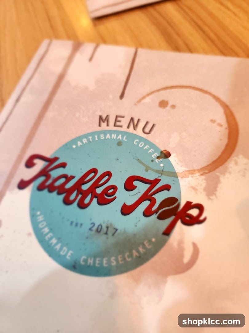 A menu with a coffee stain design. The menu is for Kaffe Kop, specializing in artisanal coffee and homemade cheesecake, established in 2017.