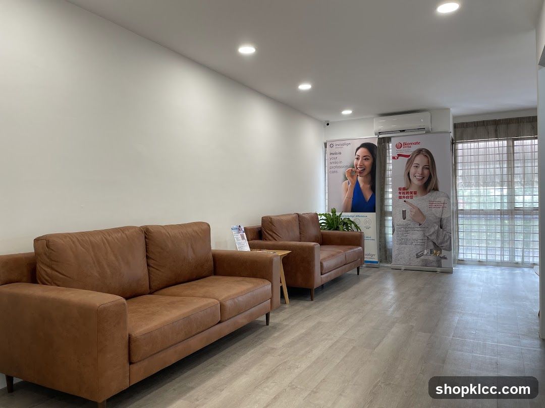 A waiting area with two brown leather sofas, a small wooden table, and two large promotional posters. One poster advertises Invisalign, and the other advertises Biomate Salign. The room has a light-colored floor and white walls, with recessed lighting. An air conditioning unit is visible on the wall above the posters. Large windows with blinds are on the right side of the room.