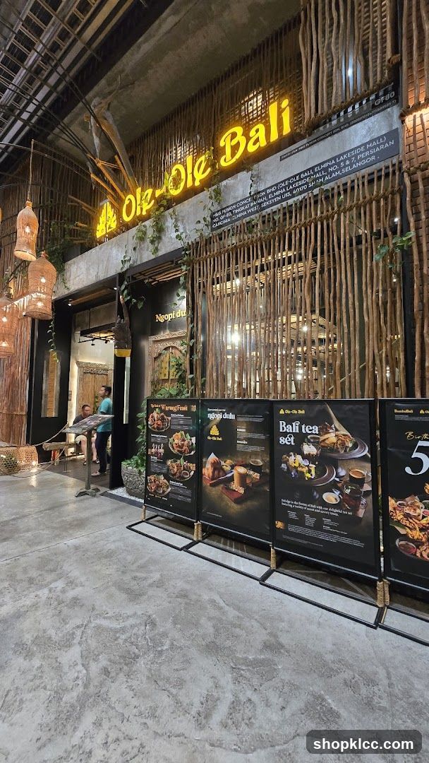 The exterior of a restaurant with a prominent neon sign that reads "Ole-Ole Bali". The entrance is decorated with natural wood elements and hanging lanterns. Several display boards with food images and text are placed in front of the establishment.