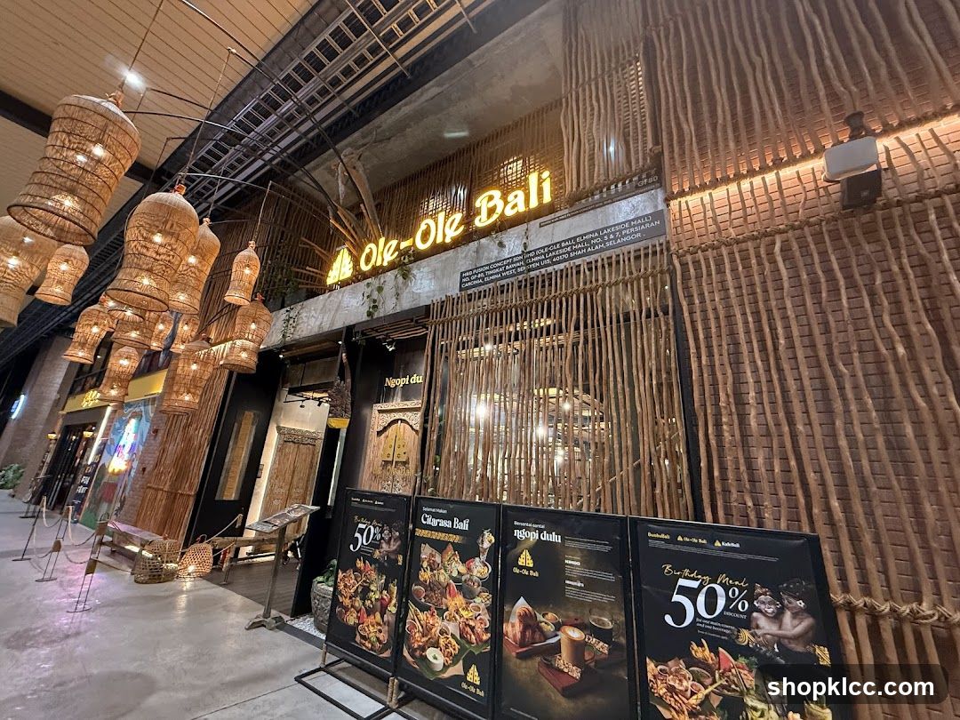 The exterior of a restaurant with a rustic design, featuring bamboo accents and woven light fixtures. The restaurant's name, "Ole-Ole Bali," is prominently displayed in neon letters above the entrance. Several promotional posters are placed in front of the establishment.