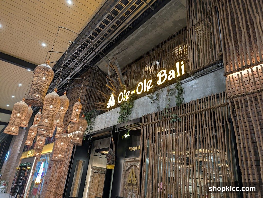 The exterior of a shop with a rustic, natural theme. The shop's name, "Ole-Ole Bali," is displayed in illuminated yellow lettering above the entrance. The facade is decorated with vertical wooden sticks and hanging woven lamps.