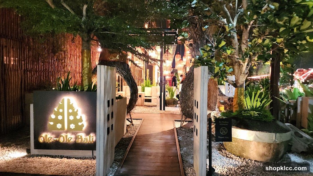 A wooden walkway leads into a restaurant with a Balinese theme. The entrance is adorned with lush greenery, bamboo fencing, and decorative lighting. A prominent sign with the restaurant's name and logo is visible on the left side.
