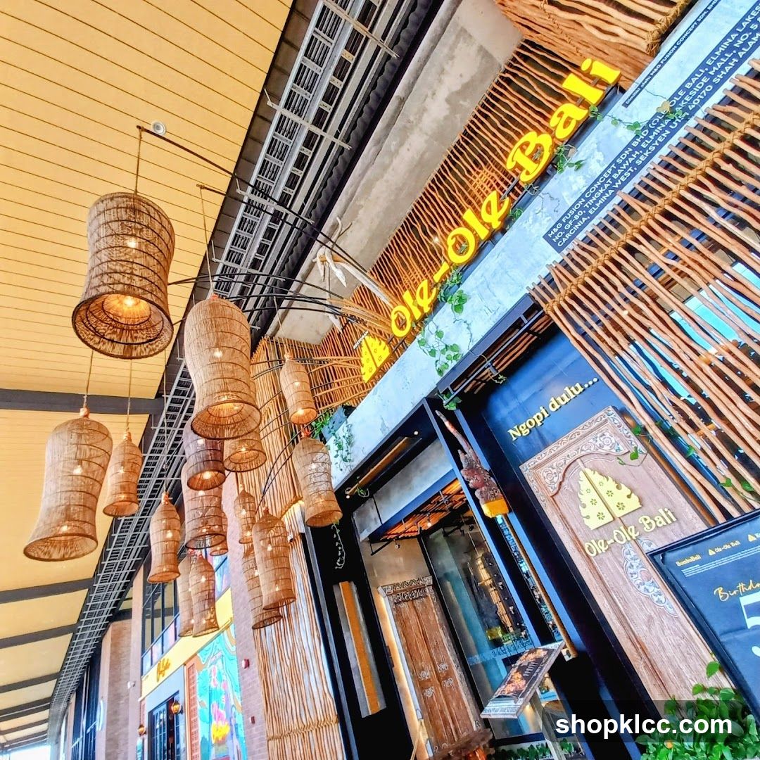 The exterior of a shop with a prominent yellow neon sign that reads "Ole-Ole Bali". The entrance features decorative wooden doors and bamboo accents. Woven pendant lights hang from the ceiling.