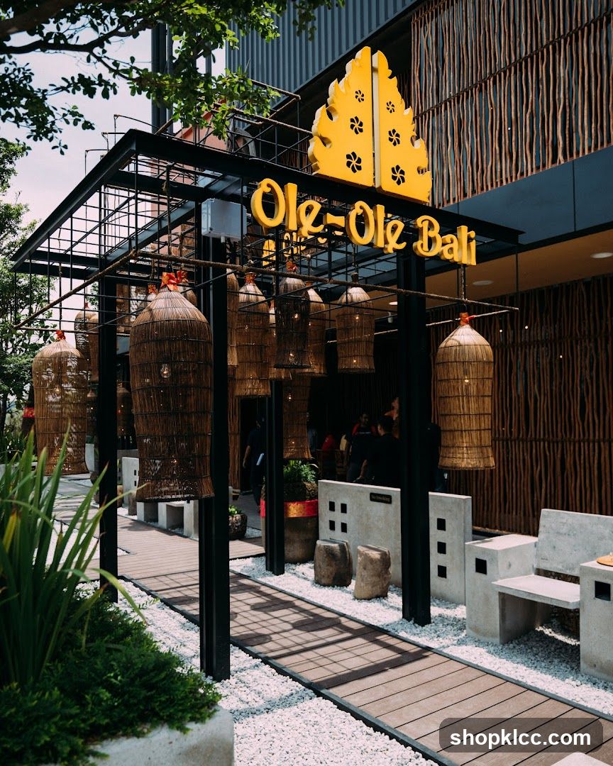 The exterior of a restaurant with a prominent yellow sign that reads "Ole-Ole Bali". The entrance features a black metal frame structure adorned with large, woven basket-like lanterns. A wooden walkway is laid out on white gravel, with concrete benches and decorative stones along the path. The building facade is covered in vertical wooden slats.