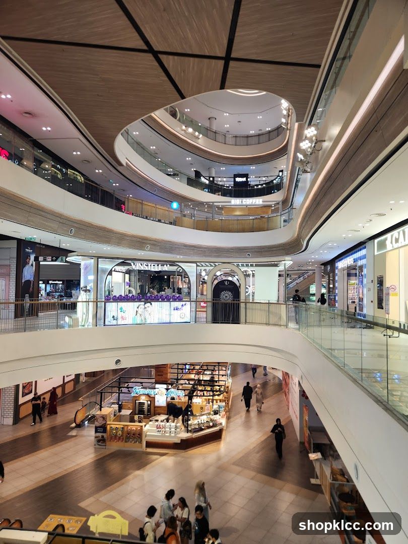 An interior view of a multi-level modern shopping mall with a contemporary architectural design. The image showcases curved balconies, wooden accents on the ceiling, and glass railings. Several shops are visible on different floors, including one with a prominent display of pastries and desserts. People are seen walking around the mall, browsing shops, and interacting. The overall atmosphere is bright and spacious.