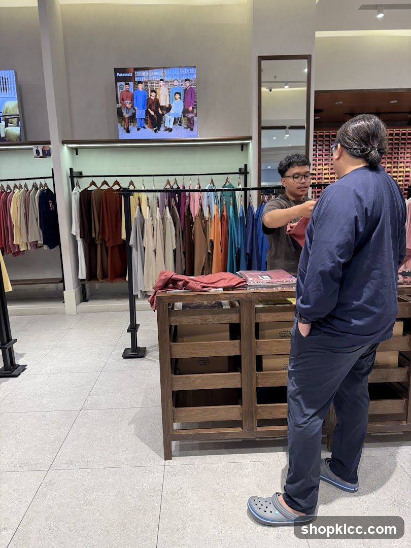 A man is being assisted by a store employee in a clothing store. Racks of colorful garments are displayed on the left, and a wooden display counter is in the foreground. A digital screen on the wall shows a group of people in traditional attire.