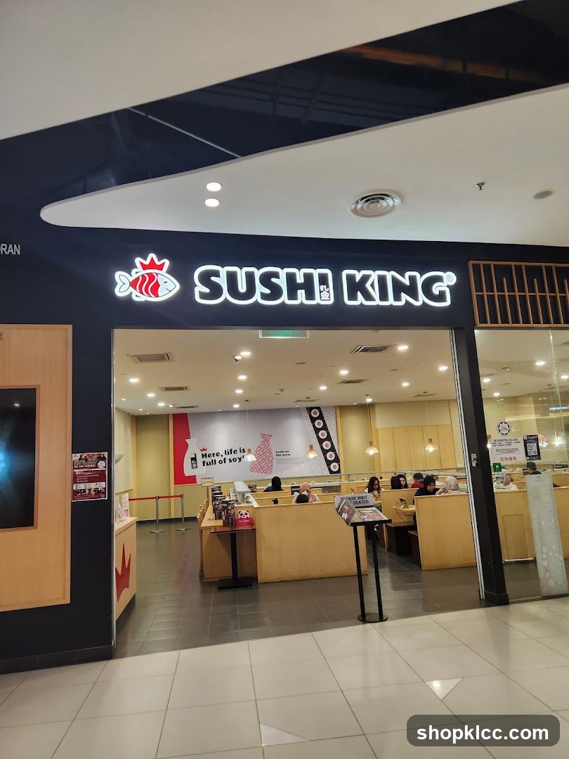 The exterior of a Sushi King restaurant. The prominent signage above the entrance reads "SUSHI KING" with a stylized fish logo. Inside, diners are seated at tables, and a conveyor belt sushi system is visible. A large banner on the back wall features the text "Here, life is full of soy!".