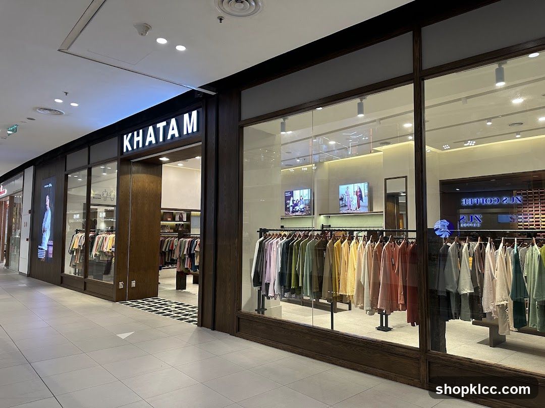 The exterior of a Khatam clothing store in a mall. The store has a dark wood facade with large glass windows displaying racks of colorful clothing. The store name "KHATAM" is illuminated in white letters above the entrance. Inside, more clothing racks and displays are visible.
