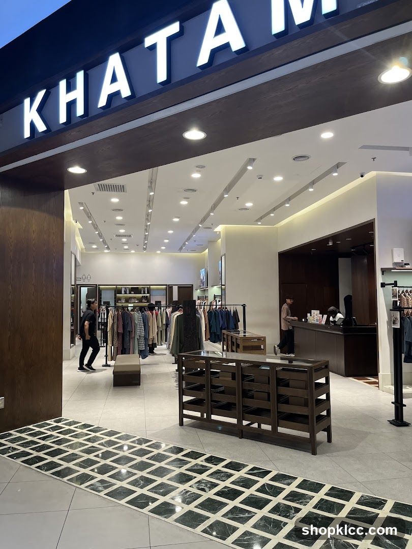 The exterior entrance of a clothing store with the name 'KHATAM' displayed in large, illuminated letters above the doorway. Inside, racks of clothing are visible, along with display shelves and a counter area where staff are attending to customers. The floor features a distinctive pattern of dark green and white tiles.