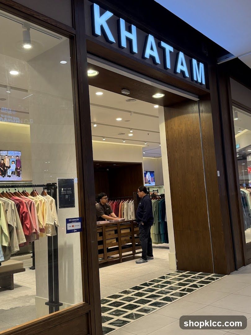 The exterior of a clothing store with the name "KHATAM" displayed in large, illuminated letters above the entrance. The entrance is framed by dark wood. Inside, racks of colorful clothing are visible, and two men are standing at a counter. A sign for "GO CASHLESS" is also visible.