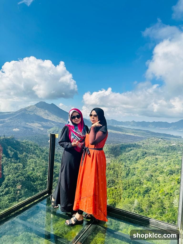 Two women posing on a glass viewing platform with a scenic mountain and lake backdrop.