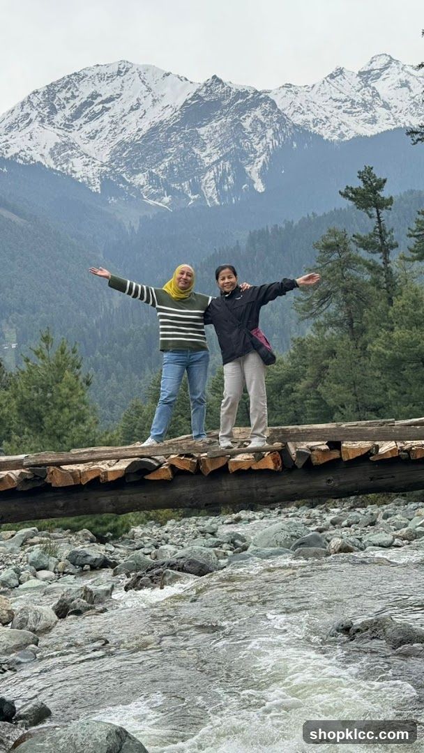Two women on a wooden bridge over a stream with snowy mountains in the background