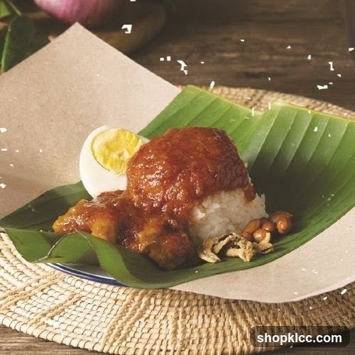 A serving of Nasi Lemak, a traditional Malaysian dish, is presented on a banana leaf. The dish includes white rice topped with a rich, reddish-brown sambal sauce, a hard-boiled egg cut in half, fried anchovies, and peanuts. The food is arranged on a woven placemat, with a piece of brown paper underneath the banana leaf. In the background, a portion of a red onion and some green leaves are visible.