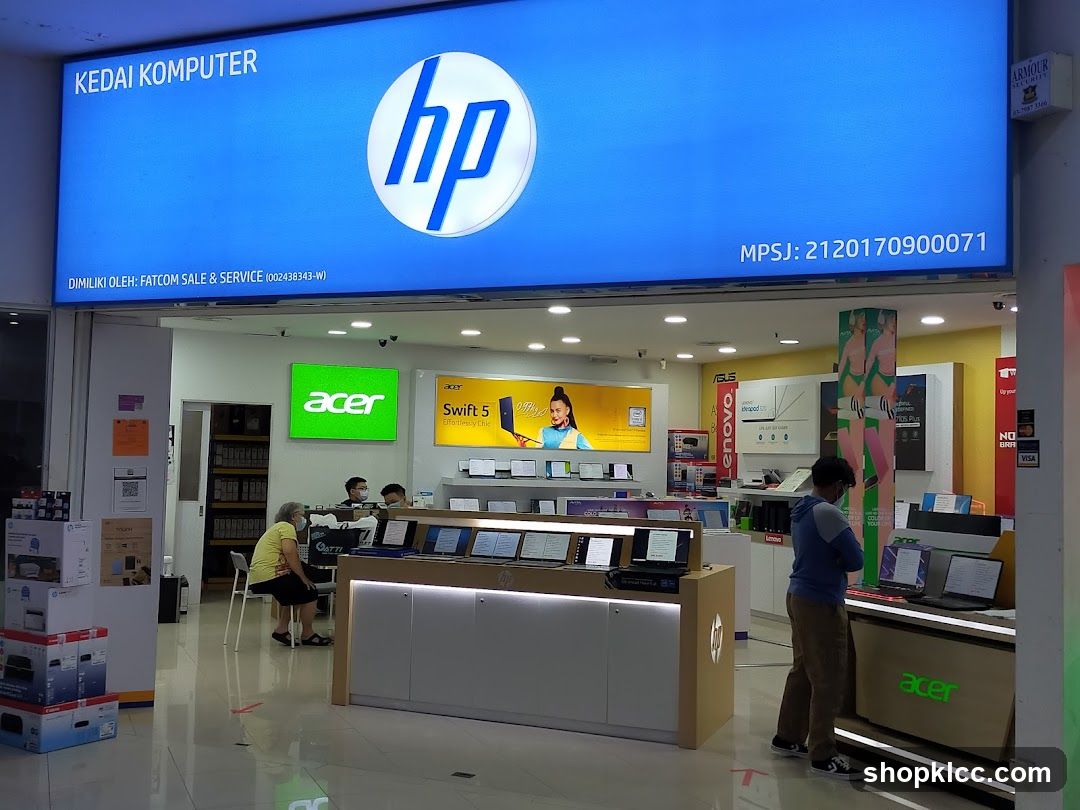 The exterior of a computer store with a large blue illuminated sign displaying "KEDAI KOMPUTER" and the HP logo. Below the sign, it states "DIMILIKI OLEH: FATCOM SALE & SERVICE". Inside the store, various computer brands like Acer, HP, Lenovo, and Asus are displayed. There are laptops on display stands, and customers are browsing.