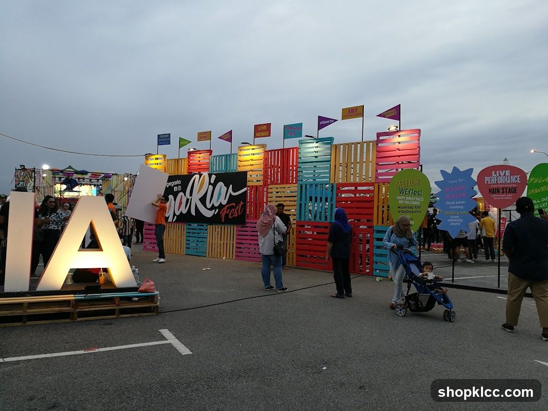 A large outdoor event features prominent illuminated letters spelling "IA" in the foreground. Behind this, a vibrant wall constructed from colorful wooden pallets serves as a backdrop. Various signs are displayed, including one that reads "Gegaria Fest" and another promoting "Selfies! We fies!". Further signs indicate "Live Performance" and "Main Stage". People are milling around the area, some pushing strollers. The sky is overcast.