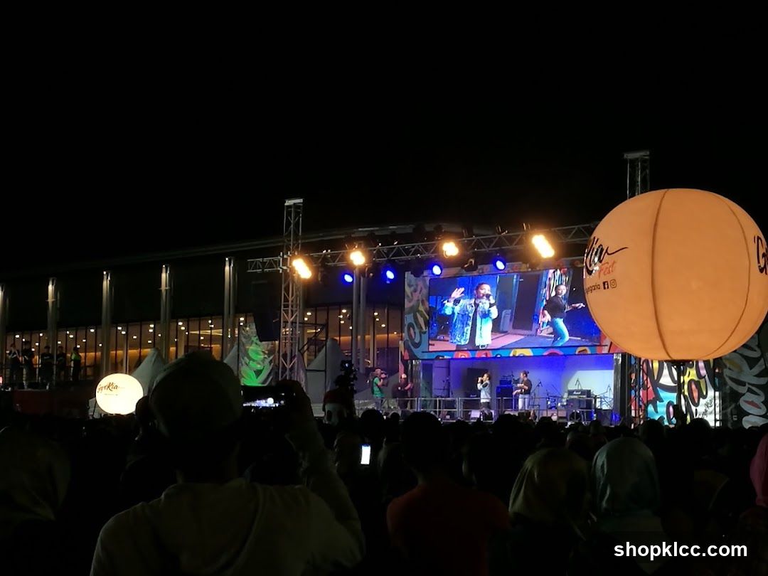 A large crowd watches a performance on a brightly lit stage at night. A large screen displays performers, and a giant illuminated balloon with a logo is visible to the right. The background shows a building with many windows and lights.