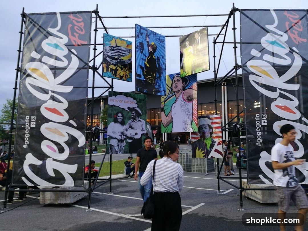 Large black banners with white text and colorful accents reading "Gegaria Fest" and "#gogegaria" flank an entrance area. Several colorful, artistic posters are hung from a metal frame between the banners, depicting various people and scenes. People are walking around the area, and a building with lights is visible in the background.