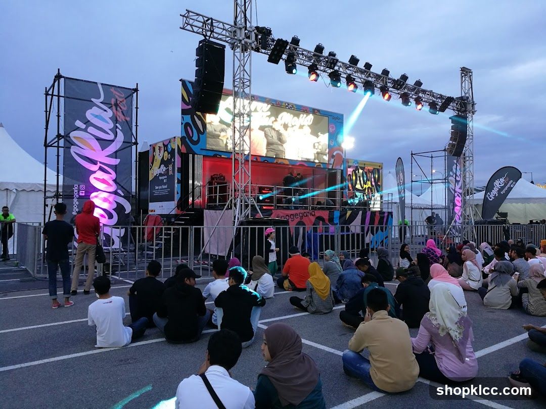 An outdoor concert or event is taking place with a stage set up. A large screen displays performers, and a lighting rig illuminates the stage. A crowd of people is seated on the ground, facing the stage, watching the performance. Banners with the hashtag #gogegaria are visible.