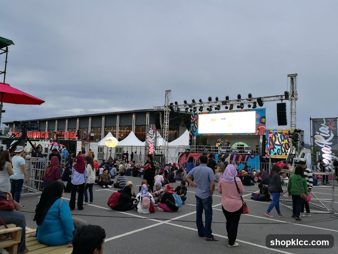 A crowd of people are gathered outdoors, sitting and standing, watching a performance on a stage. The stage has a large screen displaying graphics and lights. There are tents and structures in the background, and a large building behind them. The sky is overcast.