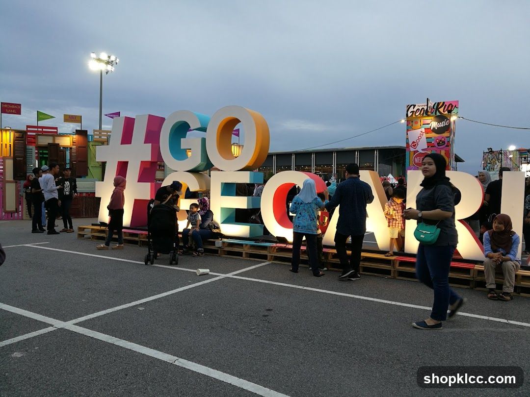 People gather around a large, colorful hashtag sign that spells out "#GO CARI". The sign is illuminated and appears to be part of an outdoor event or festival. In the background, there are various stalls and structures, including one with a sign that reads "Geal Rio Music". The sky is overcast.