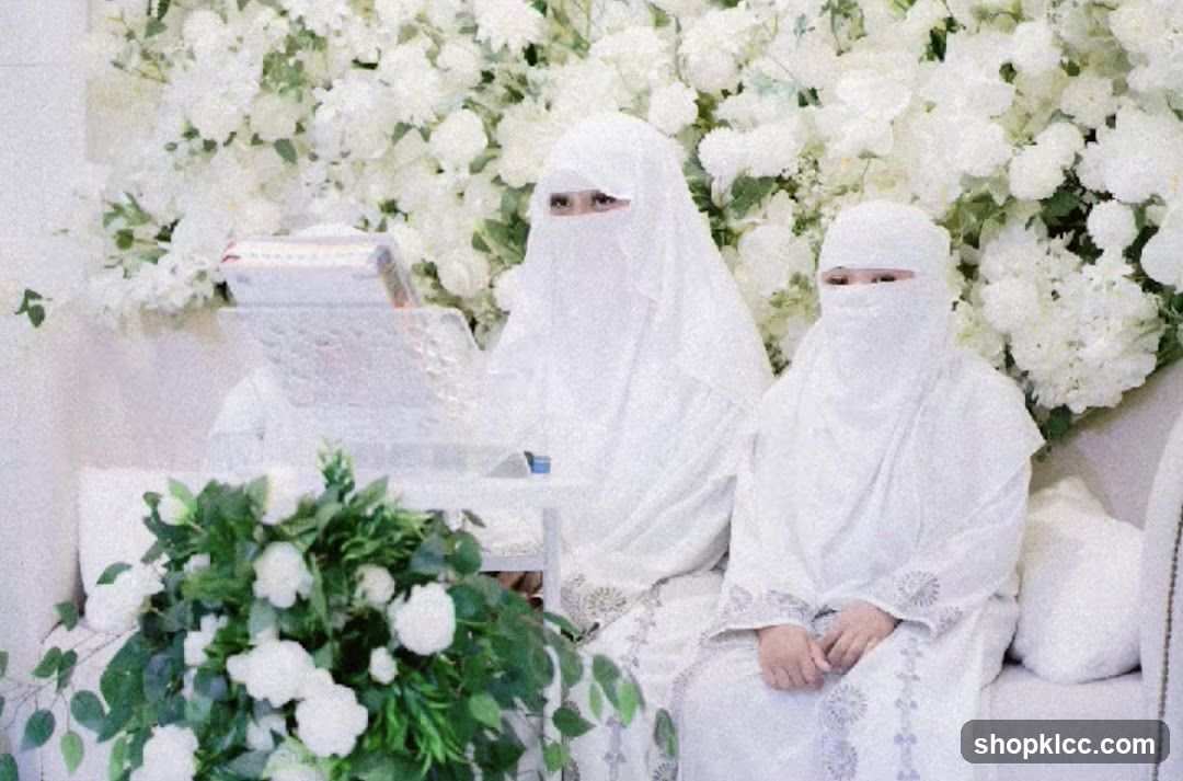 Two people in white attire with face coverings sitting in front of a floral backdrop.