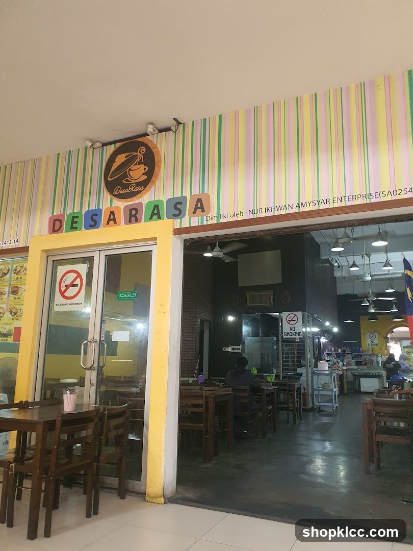 The exterior of a restaurant called Desa Rasa. The entrance has glass doors and yellow trim. Above the entrance, the restaurant name is displayed in colorful block letters and a circular logo with a coffee cup. Inside, tables and chairs are visible, along with kitchen equipment and staff.