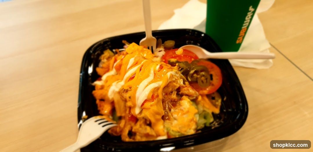 A black container filled with nachos, topped with melted cheese, sour cream, tomatoes, and jalapeños. A green Subway cup is visible in the background.