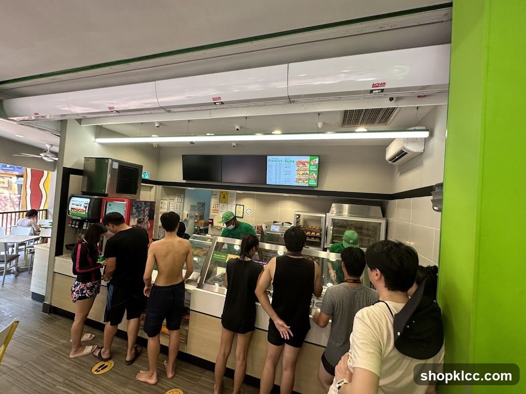 A group of people are standing in line at a sandwich shop. The shop has a bright green exterior and a counter with food displayed. A menu board is visible above the counter.
