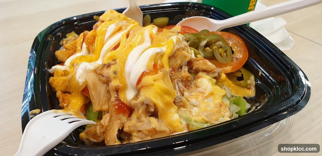 A black plastic bowl filled with a salad-like dish. The dish contains lettuce, sliced tomatoes, jalapeño peppers, and pieces of chicken. It is topped with melted cheese and a white sauce, possibly mayonnaise or ranch dressing. A white plastic fork and spoon are visible in the bowl. In the background, a green and yellow logo is partially visible, suggesting a fast-food restaurant.