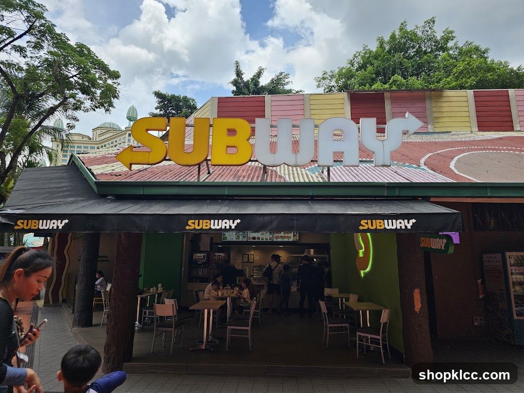 The exterior of a Subway restaurant with its prominent yellow and white signage. The storefront features a black awning with the Subway logo repeated on it. Inside, tables and chairs are visible, with customers seated and staff working. The building has a colorful, multi-paneled roof.