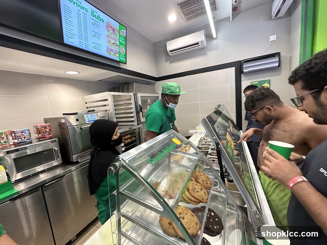 A Subway restaurant interior with customers ordering food. A digital menu board displays sandwich options and prices. Staff members in green uniforms serve customers at the counter, which features a display of cookies. Ovens and microwaves are visible in the background.