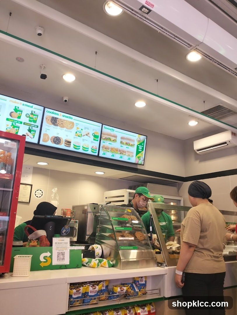 A view of the counter inside a Subway restaurant. Digital menu boards display various meal deals, cookies, snacks, and favorite sandwiches. Staff members in green uniforms are serving customers. A display case shows cookies, and shelves below are stocked with bags of chips.