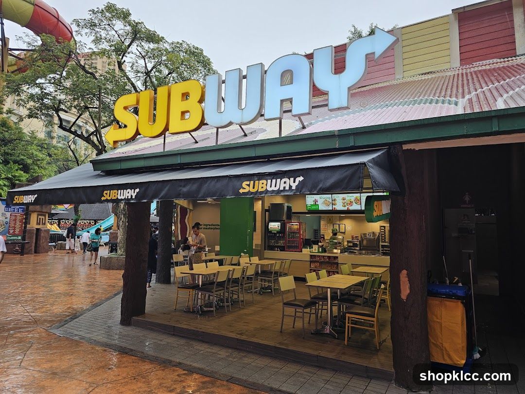 The exterior of a Subway restaurant with its prominent yellow and blue signage. The restaurant has an outdoor seating area with tables and chairs. In the background, there are elements of a water park, including a large red water slide.