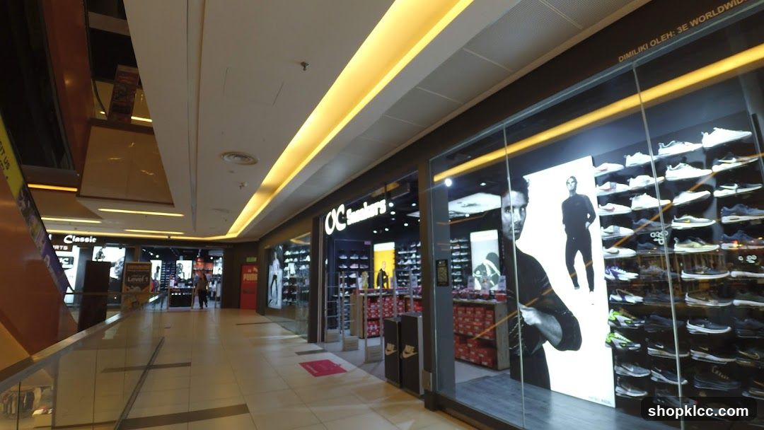 The exterior of an OC Sneakers store in a mall. The store has a large glass window displaying rows of athletic shoes. A large black and white poster of a man in athletic wear is visible on the left side of the window. The store's name, "OC Sneakers," is displayed above the entrance. Other stores, including one with a "Puma" sign, are visible in the background.