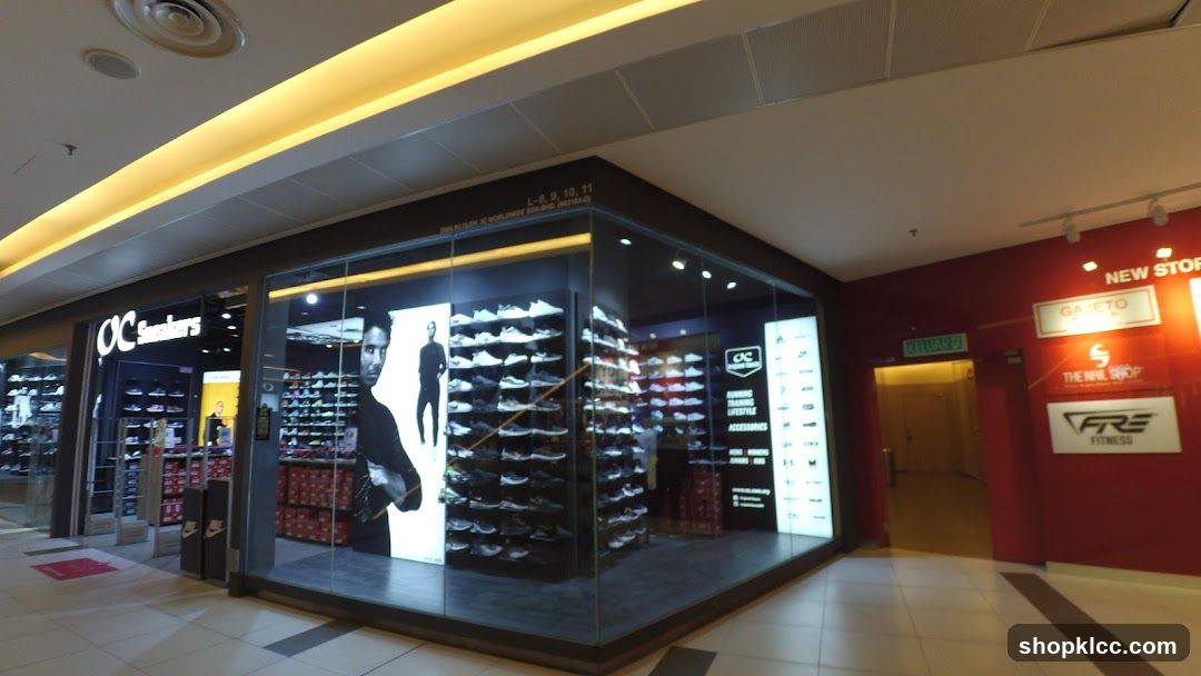A corner view of the OC Sneakers store entrance and display windows. The store features a modern design with large glass windows showcasing shelves of athletic shoes. A prominent "OC Sneakers" logo is visible above the entrance. Inside, mannequins and displays of footwear are illuminated. To the right, a red wall displays various signs for other businesses, including "NEW STORE", "GAMETO", "THE NAIL SHOP", and "FRE FITNESS".