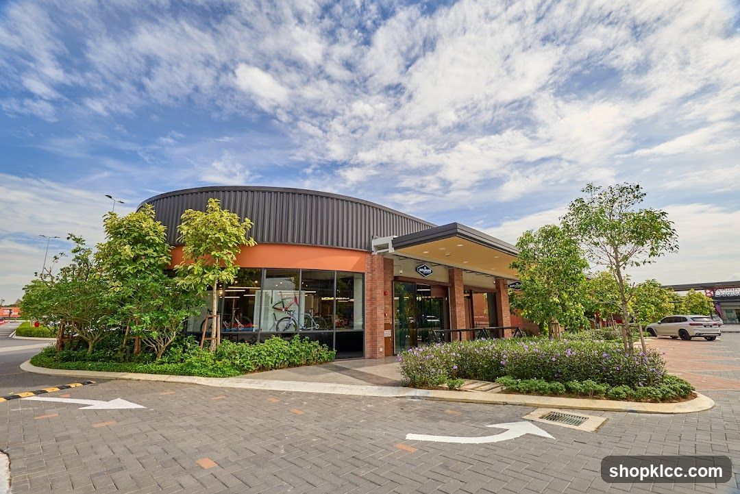 A modern bike shop with a curved roof and large glass windows displaying bicycles. The entrance is covered by an awning, and the building is surrounded by landscaping and trees. The shop's signage is visible above the entrance.
