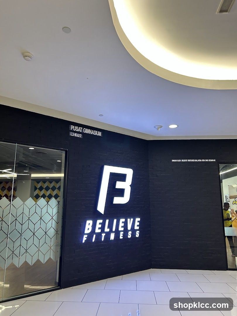 The exterior of a gym with a black brick wall and a large illuminated logo that says "B" above the words "BELIEVE FITNESS". Above the logo, text reads "PUSAT GIMNASIUM L3-16 & 17". To the left is a glass door with a geometric pattern. To the right, a person in a yellow shirt is visible through another glass entrance.