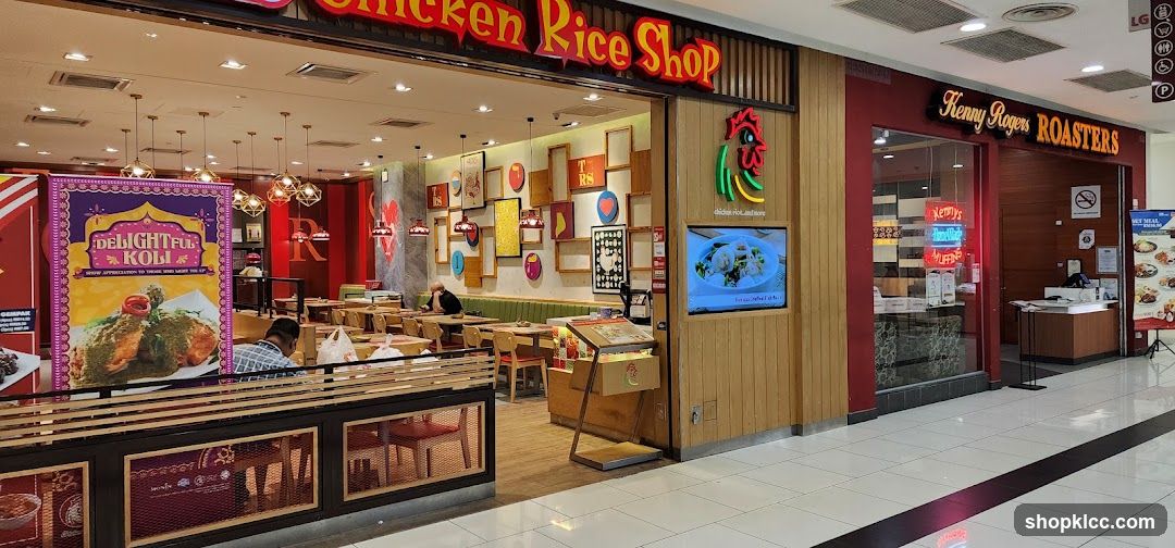A view of the storefronts for Chicken Rice Shop and Kenny Rogers Roasters, located next to each other in a mall. Chicken Rice Shop has a prominent sign with red lettering and a chicken logo. Inside, tables and chairs are visible, along with decorative wall art. Kenny Rogers Roasters has its own sign and a counter area. A promotional banner for "Delightful Koli" is displayed in front of Chicken Rice Shop.