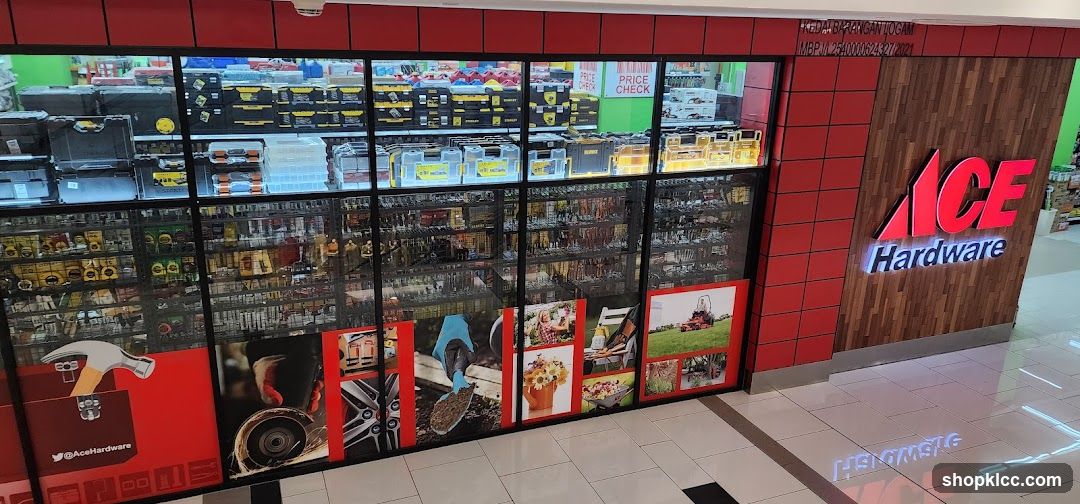 The exterior of an Ace Hardware store is visible, featuring a large display window showcasing various tools and hardware items. The store's prominent red and white logo, "ACE Hardware," is displayed on a wooden panel to the right of the entrance. The window is divided into sections, with the upper part displaying toolboxes and the lower part featuring images of gardening and DIY projects.