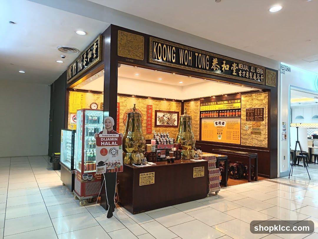 The exterior of a Koong Woh Tong shop, specializing in herbal jelly. The shop has a traditional wooden facade with ornate gold lettering and decorative elements. A large sign above the entrance reads "KOONG WOH TONG" and "MENJUAL JELI HERBA". Inside, shelves are stocked with bottles of herbal jelly, and there are displays of traditional tea urns. A refrigerated display case is visible to the left, containing various beverages. A cardboard cutout of a woman in a headscarf holding a sign that says "PRODUK KAMI DIJAMIN HALAL" (Our products are guaranteed Halal) stands in front of the display case. A menu board is mounted on the wall inside the shop.