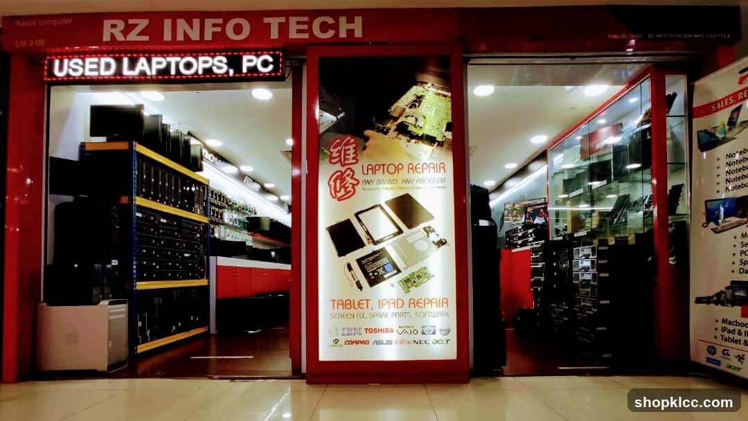 The storefront of RZ INFO TECH, a computer repair and sales shop. The shop has a red facade with the business name prominently displayed. Inside, shelves are filled with computers and laptops. A large illuminated sign advertises laptop and tablet repair services, listing brands like IBM, Toshiba, Sony, Compaq, Asus, and NEC. Another sign indicates they sell used laptops and PCs.