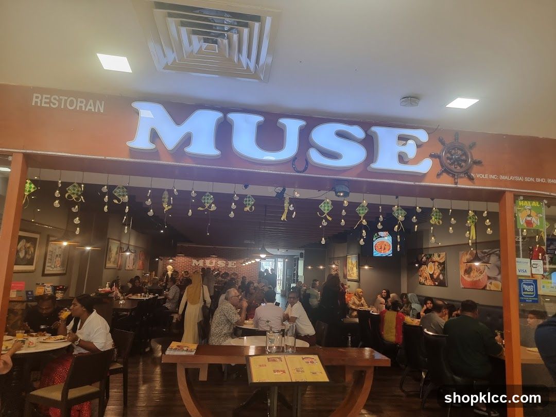 The exterior of a restaurant called Muse, with large illuminated letters spelling out the name. The restaurant appears to be busy with patrons dining inside. Decorative elements, possibly for a festival, hang from the ceiling.