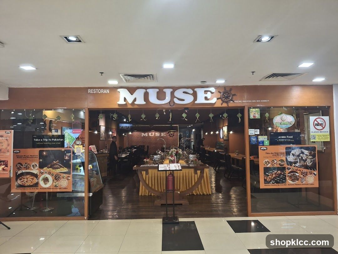 The exterior of a restaurant called Muse, with large illuminated letters spelling out the name above the entrance. The restaurant has a warm, inviting interior visible through the glass facade, featuring wooden tables and chairs, and brick walls. Promotional posters showcasing food are displayed on the left and right sides of the entrance.